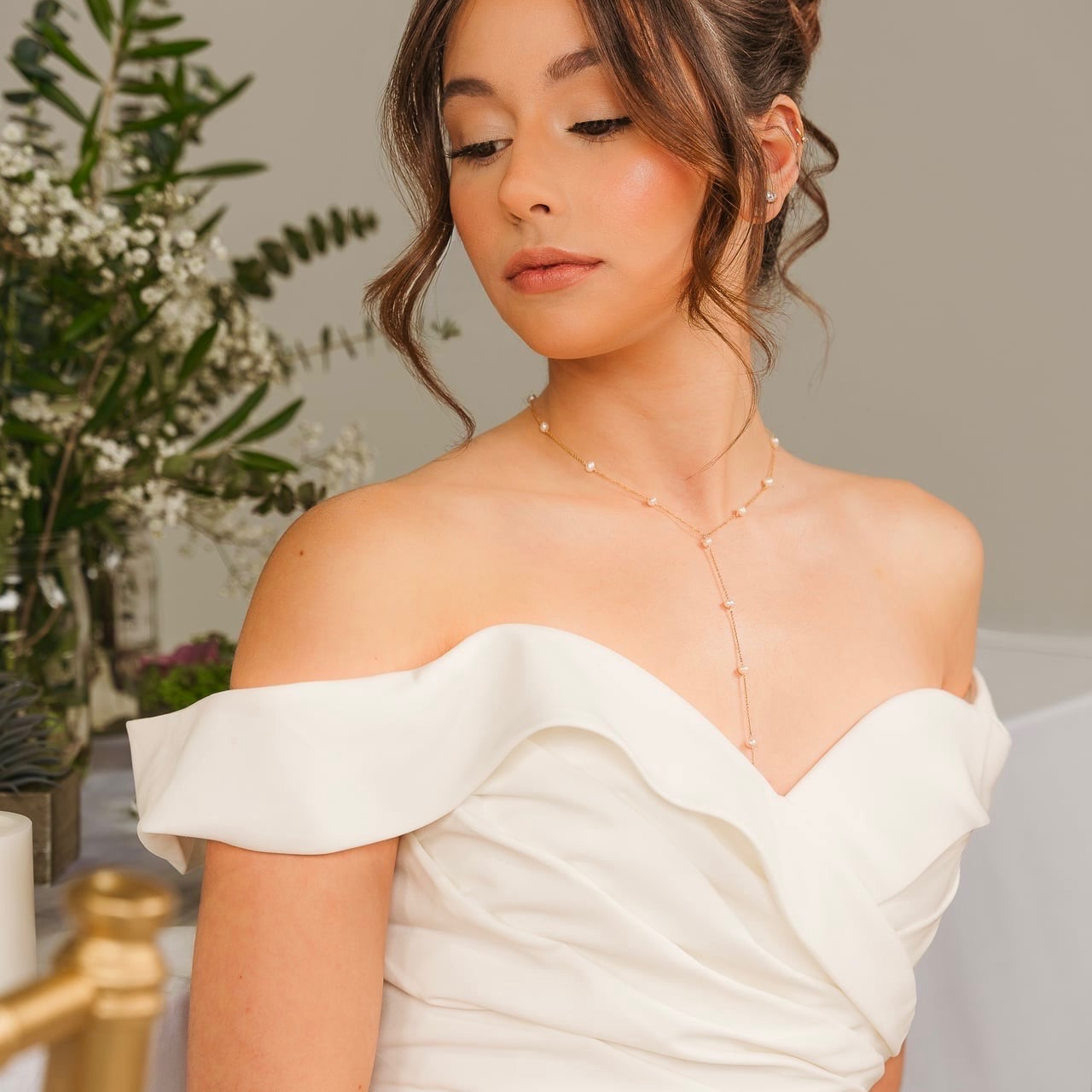 Woman in a white off-shoulder dress with floral decorations in the background with permanent necklace from Lavish Everlasting